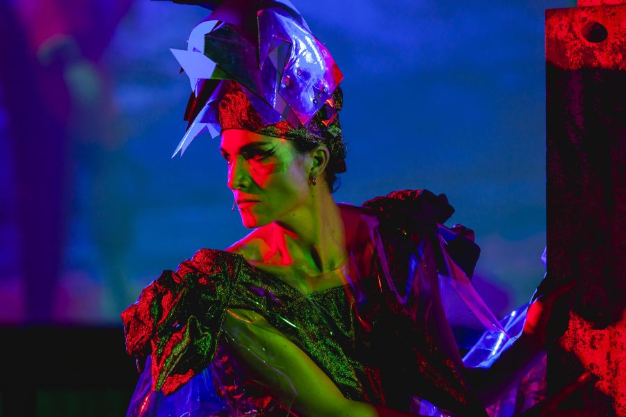 A woman artist (Justene Williams) photographed performing onstage under neon coloured lights.