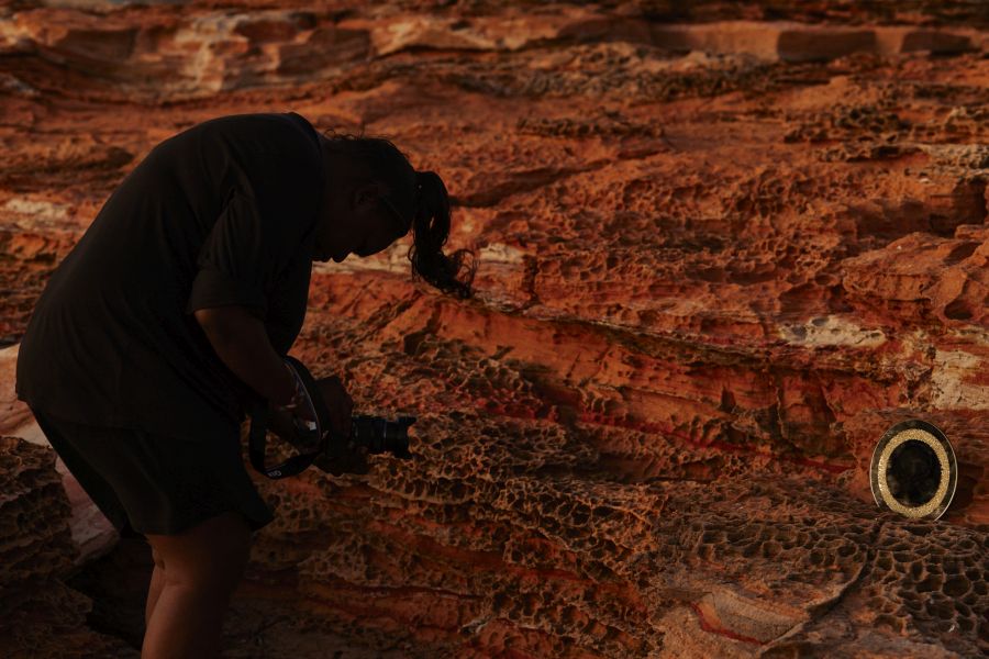 a shadow of an artist crouching over a camera against a red earth rocky outcrop in the Kimberley region of Australia.