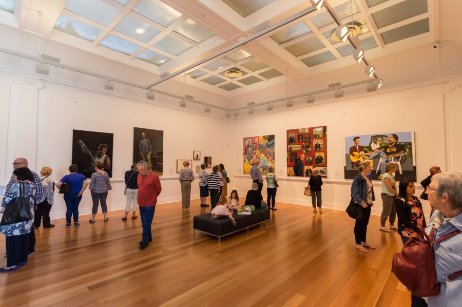 Audiences roaming around a gallery space looking at contemporary portrait paintings.