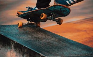 A close up photo of a skateboarder's deck: all we can see is the board and its wheels angled against a ramp and a glimpse of the person's shoes.