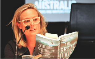 A blonde-haired white woman wearing glasses reads from a novel at a festival reading.
