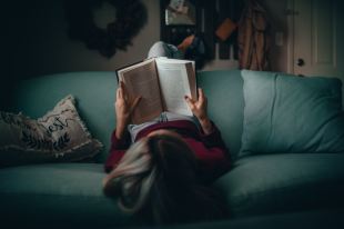 A woman in a red shirt sits on a couch reading a book. She is photographed from above so that we are looking down at the top of her head and the book in her hands.