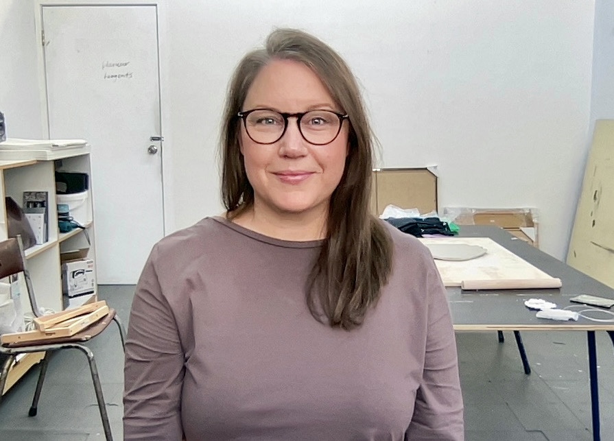 A woman with long brown hair and glasses smiles at camera. A desk and chairs are behind her.