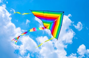 A triangular rainbow-coloured kite flies against a blue, cloud-dotted sky.