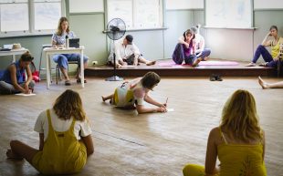 A group of people in a rehearsal room with timbre floor sitting in a loose circle. There is a women sitting and learning her body down to write on a piece of paper in the centre of the image.