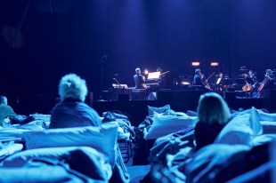 People in camp beds, photographed from behind, watch an ensemble play a 'lullaby for grown-ups' on stage.