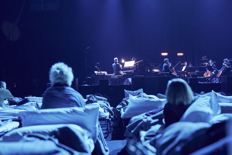 People in camp beds, photographed from behind, watch an ensemble play a 'lullaby for grown-ups' on stage.