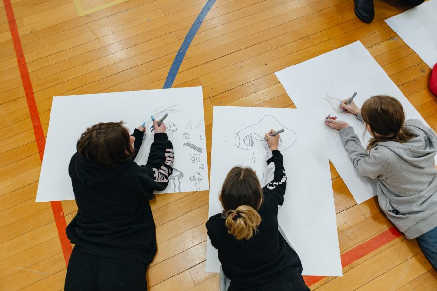 Three high school students lying on the floor drawing on large piece of white paper.