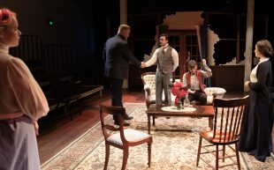 A theatre set with five actors on stage. The set depicts a living room interior with patterned carpet, a lounge sofa, table and two chairs. In the centre are two men shaking hands, next to them is a women sitting on the sofa. Two female figures stand to either side.