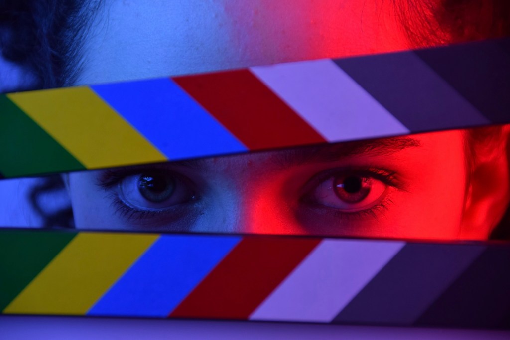 arts news. Image is a woman's eyes peering through a raised colourful clapperboard.