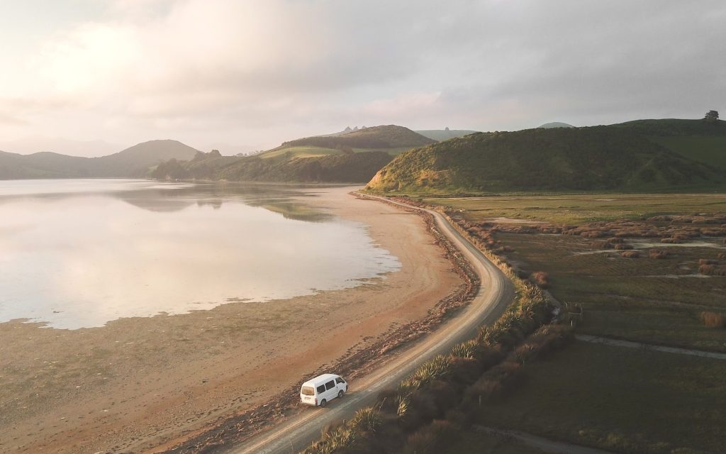 A sparse landscape with a body of water on the left and green plains on the right. In the middle is a road with a small van. The sky is grey and gloomy.