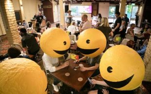 A crowded pub where four figures with big smiley face headwear sit near a table.