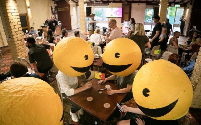 A crowded pub where four figures with big smiley face headwear sit near a table.