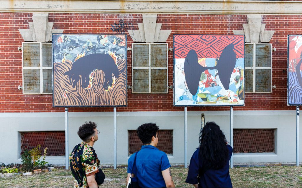 Panels of artworks installed outside a redbrick building. The works depict colourful First Nations motifs. Three figures are standing in front of the panels looking at them.