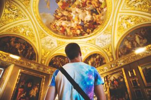 A man wearing a multi-coloured t-shirt stares up at a gilded ceiling, frescos and paintings in Florence, Italy.