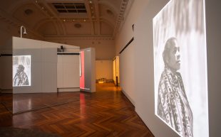 The inside of a heritage building with polished wooden floor and high curved ceiling. In the space is a woman projected onto the right wall, and a mirrored surface on the left side reflecting the projection.