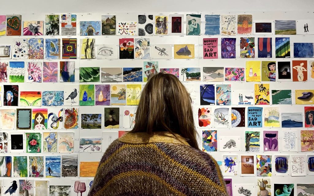 A wall filled with colourful images in A5 size. Standing in front is a person with long brown hair and a brown jacket.