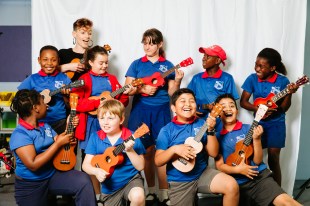 A group of school children with ukeleles are laughing. They are accompanied by a teacher.