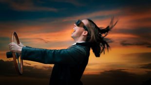 A white woman wearing motoring goggles holds a steering wheel. Her head is thrown back, her long hair blowing in the wind, and she is laughing or shouting exuberantly.