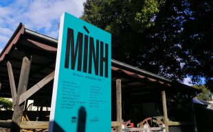 Teal coloured promotional sign with word MÌNH in bold capital letters, in front of trees.