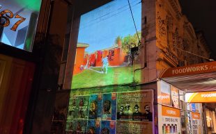 Performance video projected onto a red brick wall at the side of the street with shops. A performer in an otherworldly silver and fluid costume moving and interacting around a highly and vividly decorated rotating clothes line, filled with vibrant fabrics in a suburban Australian home, with a green garden and wooden fence at the dusk of sunset.
