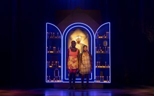 Two young Black women stand on stage in front of an open panel with illuminated blue and gold light. In the centre of the panel is a male figure with gold light illumination around him.