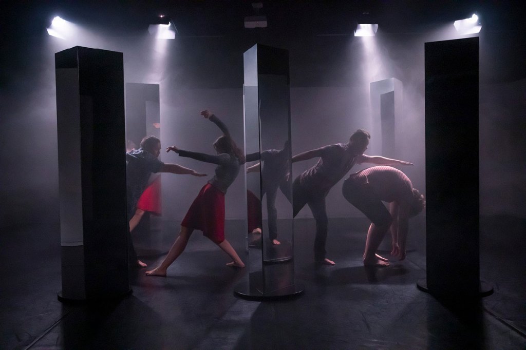 Four performers dance between mirror plinths in a gloomy space