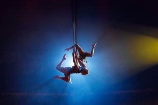 Two young men perform an aerial straps act, silhouetted against a bright light.