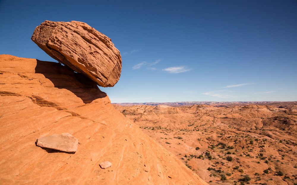 A sandstone boulder tilts dangerously on the top of the hill. In the background is the sky and a earthy, barren landscape.