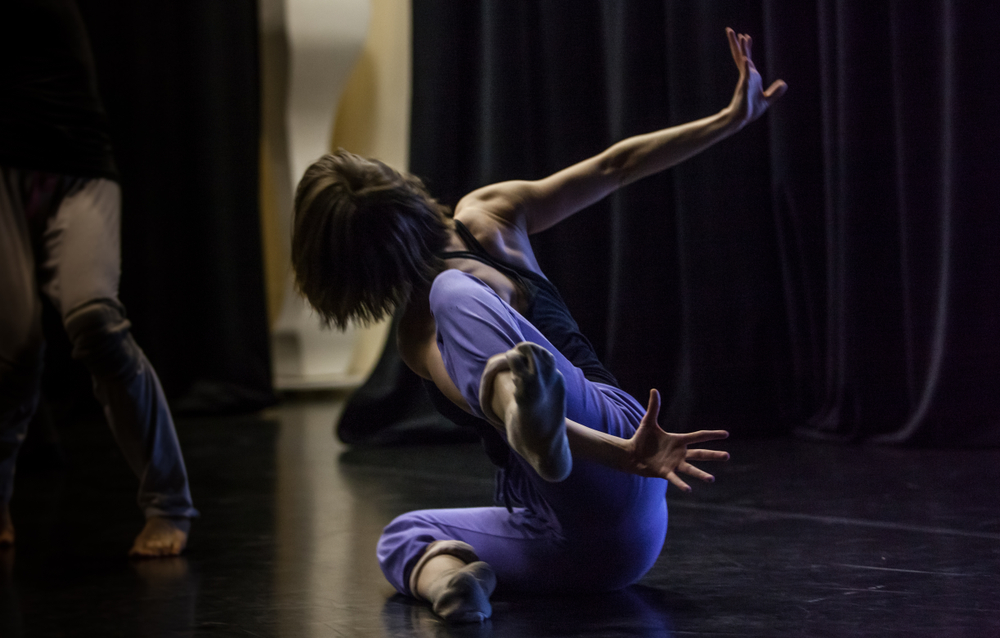 A contemporary dancer caught-mid movement on the studio floor. Her arms are outstretched behind her, her left leg looped over her left arm in a dynamic pose.