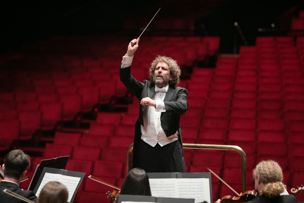 A white man in evening dress with baton raised dramatically conducts an orchestra, who are barely visible. The empty seats behind him suggest this is a rehearsal photo.
