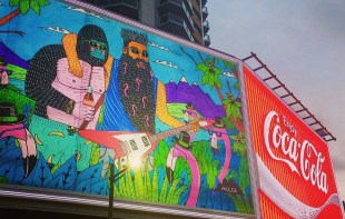 a colourful graphic style mural of a gorilla and a bearded person in nature, displayed as a giant billboard next to a Coca Cola billboard advertisement.