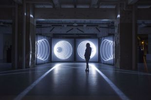 A darkened warehouse space with a single female figure looking at a bright white light art installation that is the room's only light source.