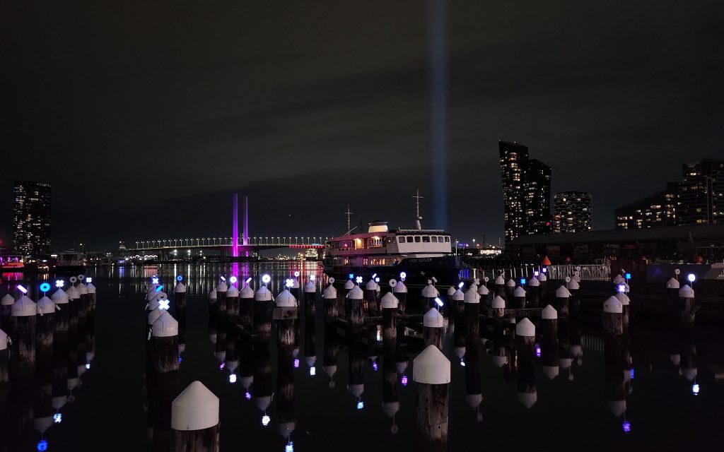 Pier and city scape on a cold winter evening. Light installations on top of poles in the water, with a blue light beam in the background.
