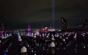 Pier and city scape on a cold winter evening. Light installations on top of poles in the water, with a blue light beam in the background.
