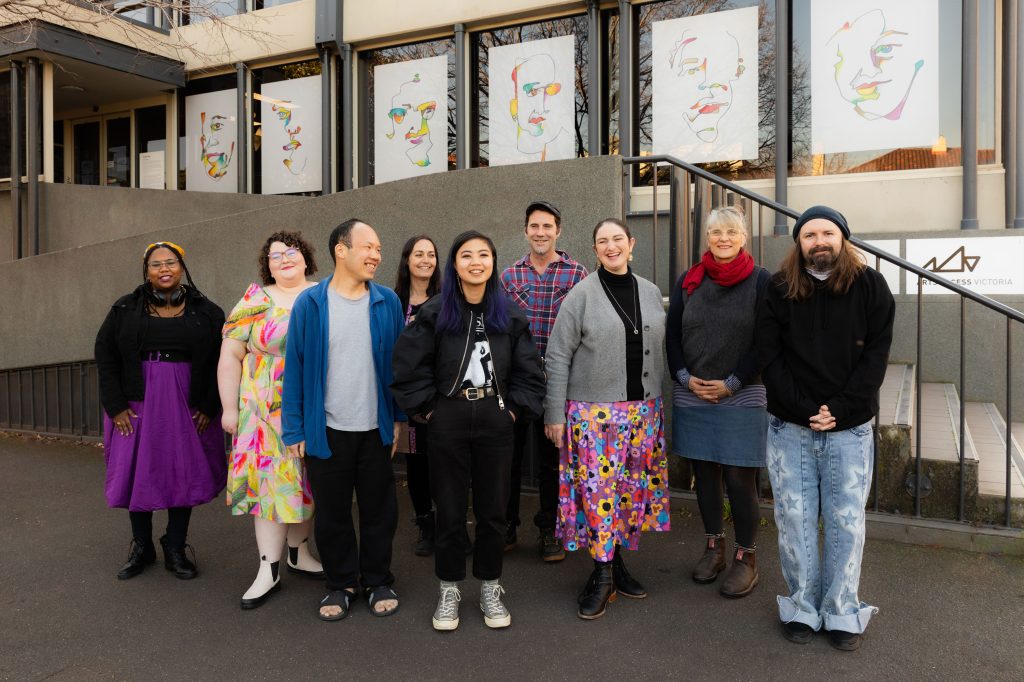 Line-up of nine people standing in front of Arts Access Victoria's office, each window displaying art images of faces