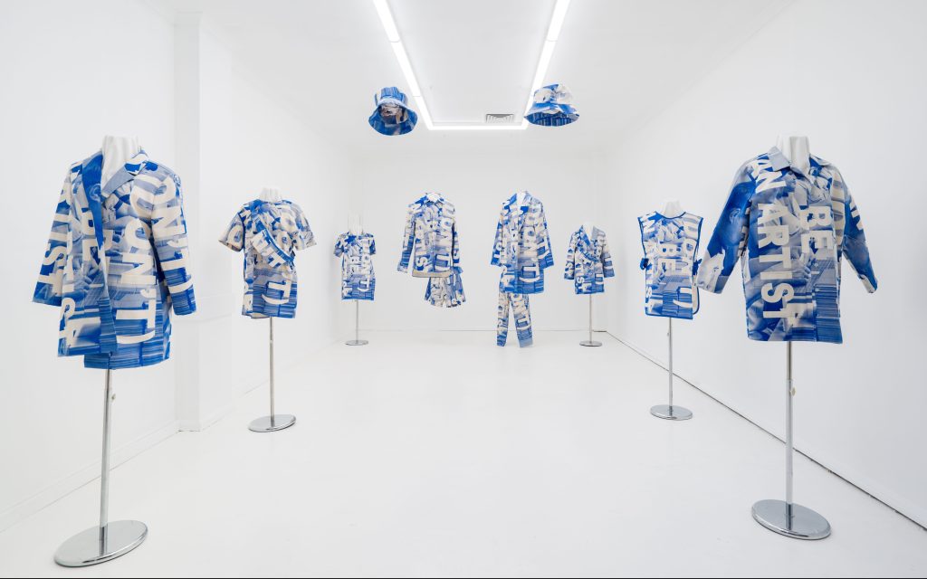 A series of blue and white screen printed workers uniforms, arranged like a shopfront display. They feature the writing '(DON'T) BE AN ARTIST' in a newsprint style.
