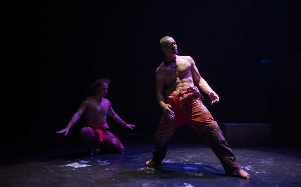 Two male dancers, wearing orange prison overalls rolled down to the waist. Their chests and arms and the floor are streaked with white ochre. Chandler Connell is to the left, in the background, kneeling, arms outstretched to his side. Alexander Abbot is to the right in the foreground. He is standing, legs wide, and leaning back. The background is black.
