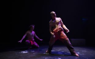 Two male dancers, wearing orange prison overalls rolled down to the waist. Their chests and arms and the floor are streaked with white ochre. Chandler Connell is to the left, in the background, kneeling, arms outstretched to his side. Alexander Abbot is to the right in the foreground. He is standing, legs wide, and leaning back. The background is black.