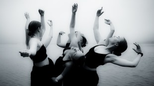 A black and white image of six figures dancing in a circle. Their arms are outstretched as some of them bend backwards and others inwards. They are wearing black crop tops.