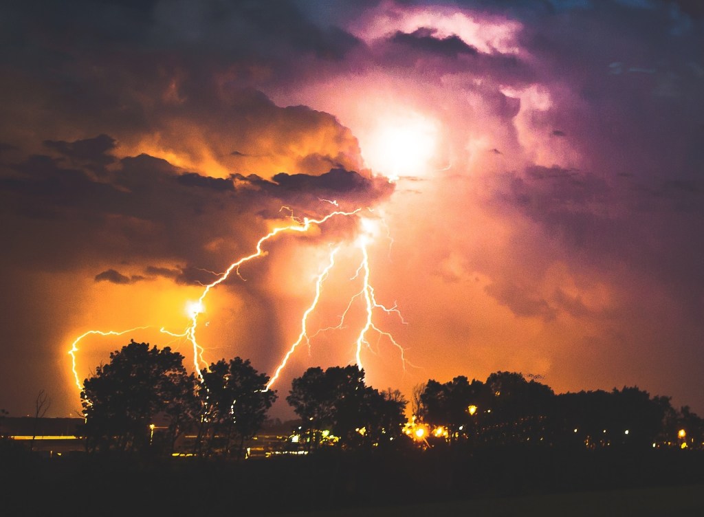 A landscape scene of stormy clouds with lighting striking some trees and buildings.