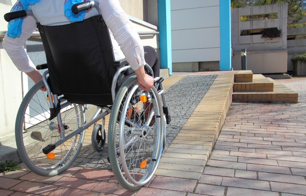 A low-angle photograph of a woman in a wheelchair at the bottom of a wheelchair ramp providing access to a building.