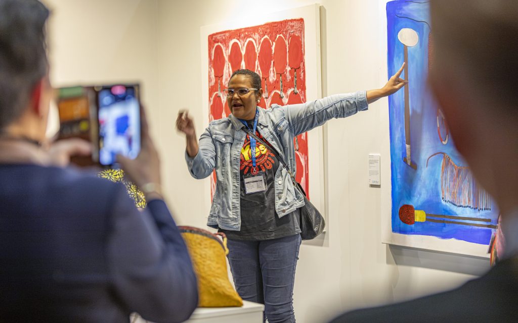 CIAF. A woman in a Vote 'Yes' T-shirt, grey denim jacket and darker grey trousers, stands in front of an orange and white painting and points at a blue painting, while a man with his back to us films her on his phone camera.
