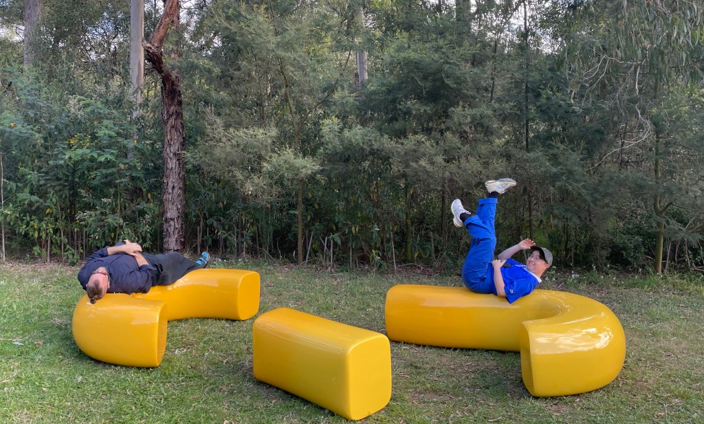 Two people sitting playfully on yellow bench in parkland. 