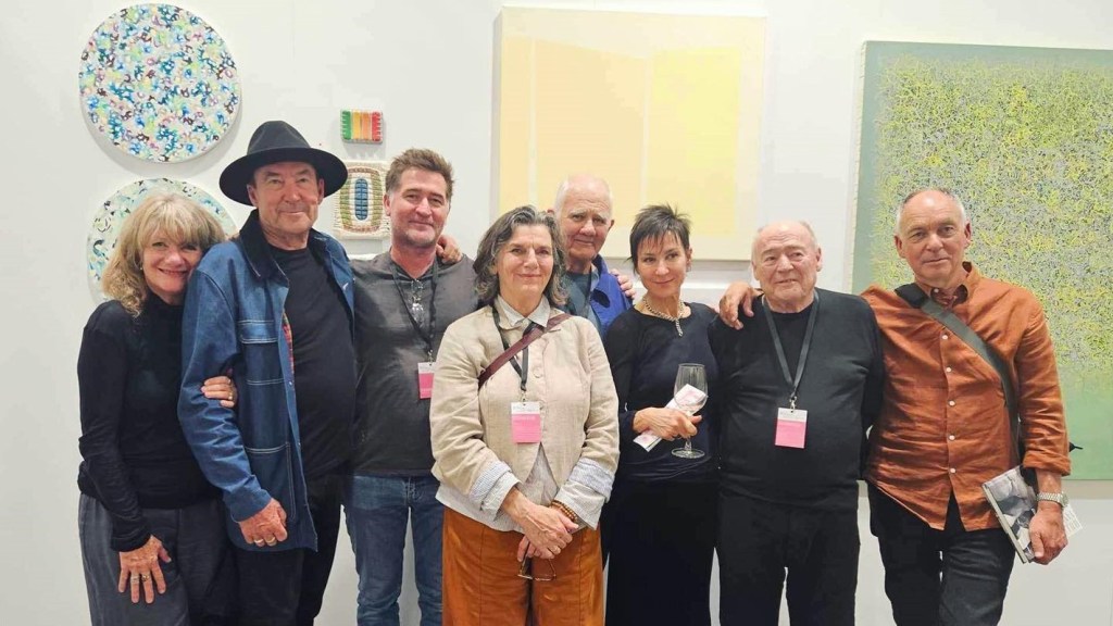 Eight Art Collective WA artists ranging in age from around sixty to eighty, posing for a photo in front of their work in a gallery space.