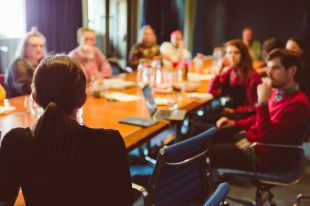 A group of arts workers sit around a conference table, intently focused on a speaker whose back is towards the camera.