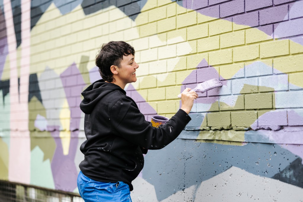 Artist insurance. Photo of a person wearing a black hoodie and blue jeans, smiling as they paint a colourful mural with a paint brush. In their other hand they are holding a paint bucket.