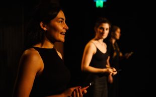 WAAPA’s Bachelor of Performing Arts (Honours). Two young woman are on stage performing and wearing sleeveless black tops. A third figure is in the background.
