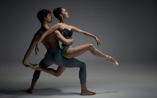 Photo of a male ballet dancer holding up a female ballet dancer against a dark grey background.