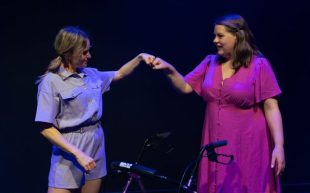Betsy and I. Two figures fist-bumping each other on a darkened stage. The figure on the right is a woman with brown hair and wearing a purple dress. The figure on the right is a woman with brown pigtails wearing a light purple jumpsuit.